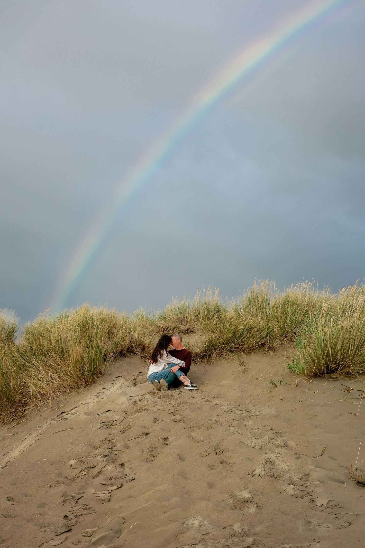 A rainbow arches over sandy dunes where people are resting in the beach grass.