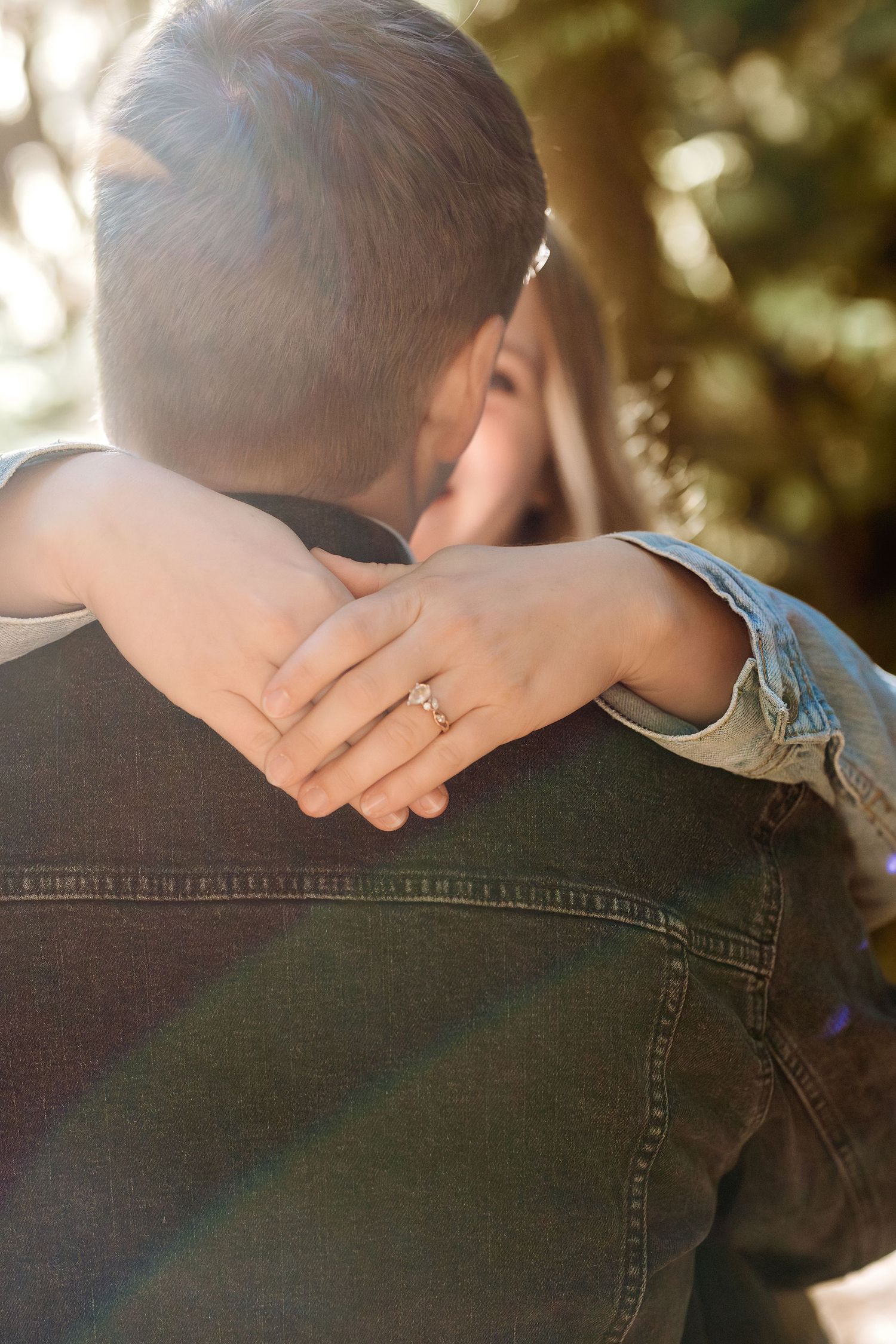 Close-up view of hands embracing with a diamond ring visible against denim clothing in natural sunlight.