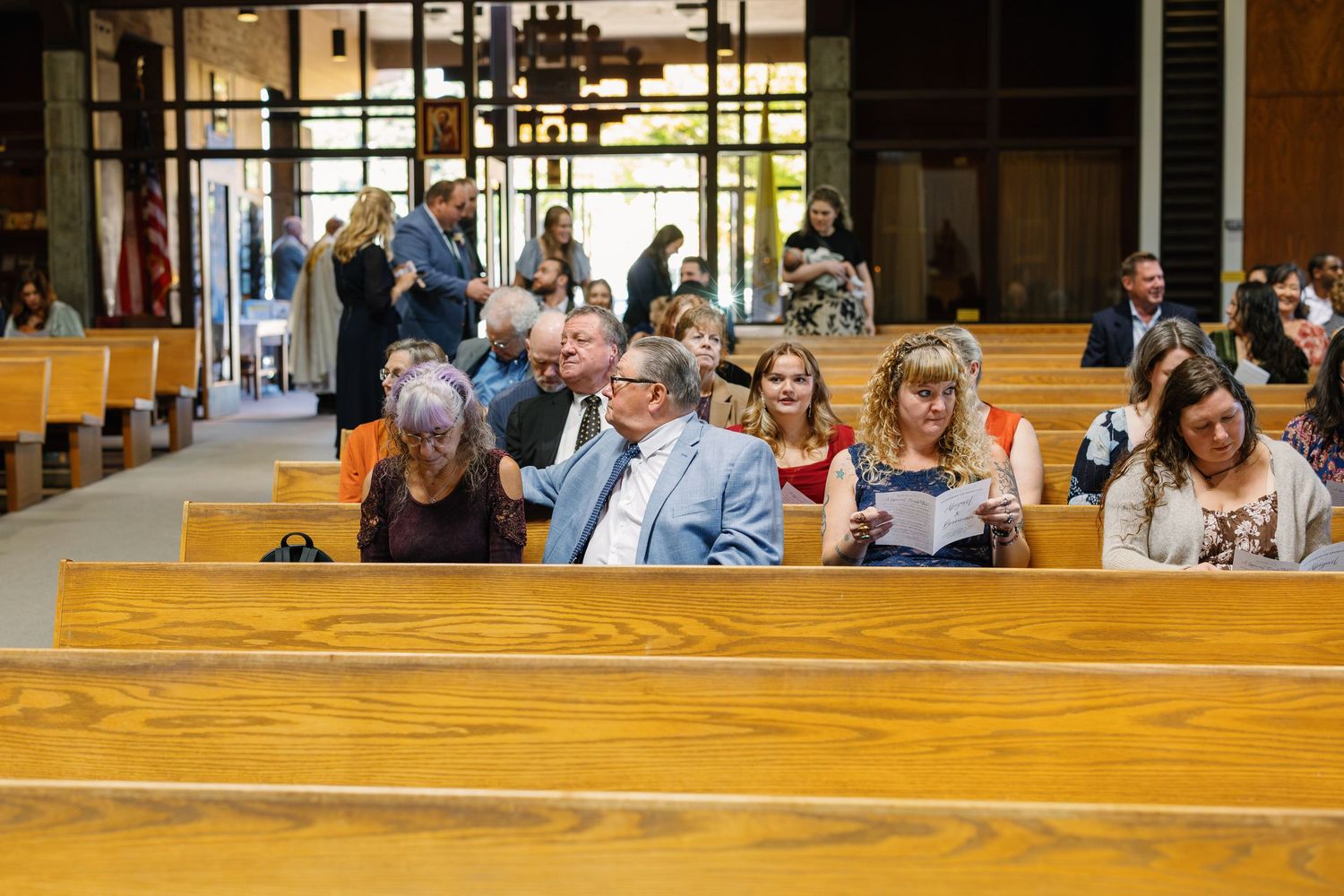 Wedding ceremony in a church with wooden pews and sunlight streaming through stained glass windows.