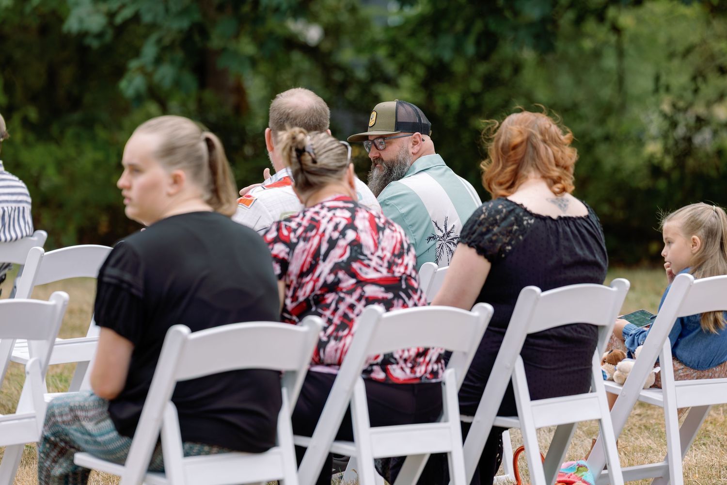 Wedding guests seated in white chairs during an outdoor ceremony surrounded by greenery and trees.