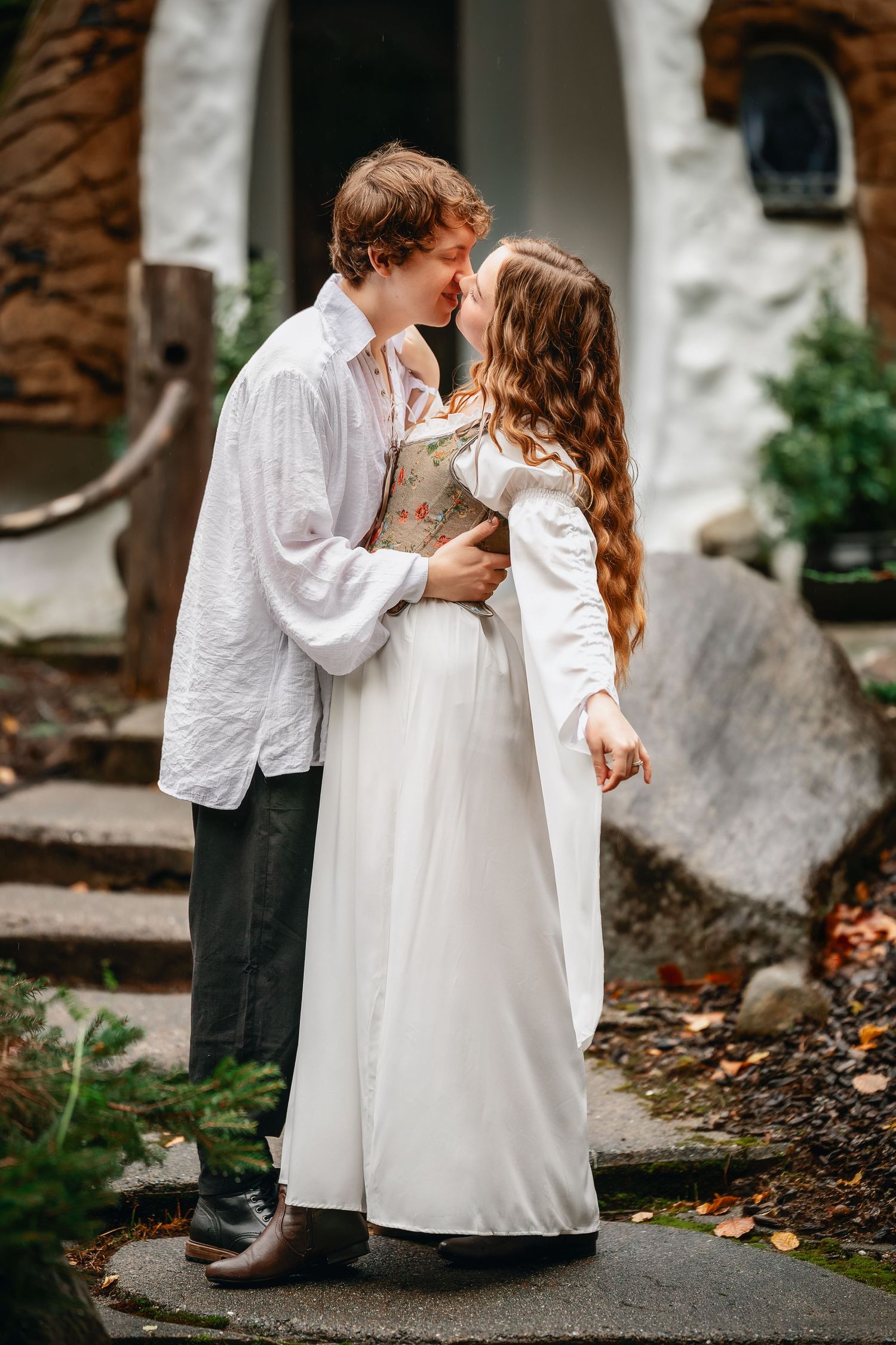A couple in vintage-inspired white clothing share a romantic moment on stone steps outside a historic building.