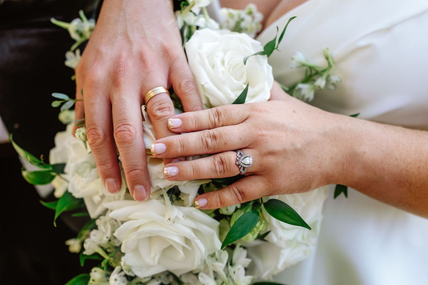 Hands wearing wedding rings rest on a white rose bouquet with green foliage.
