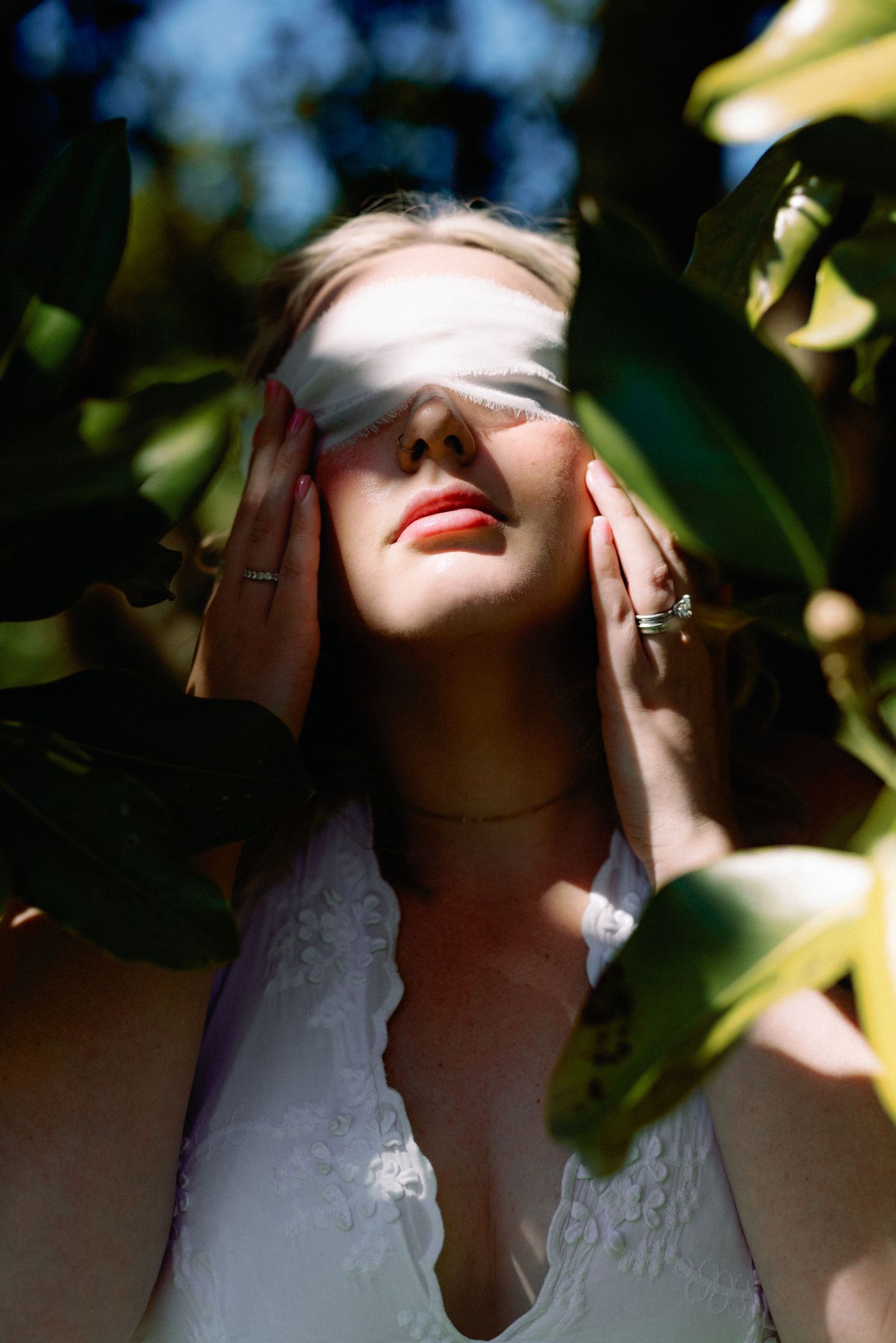 Bride in white lace dress surrounded by green leaves, hands on face with sunlight casting shadows across her eyes.