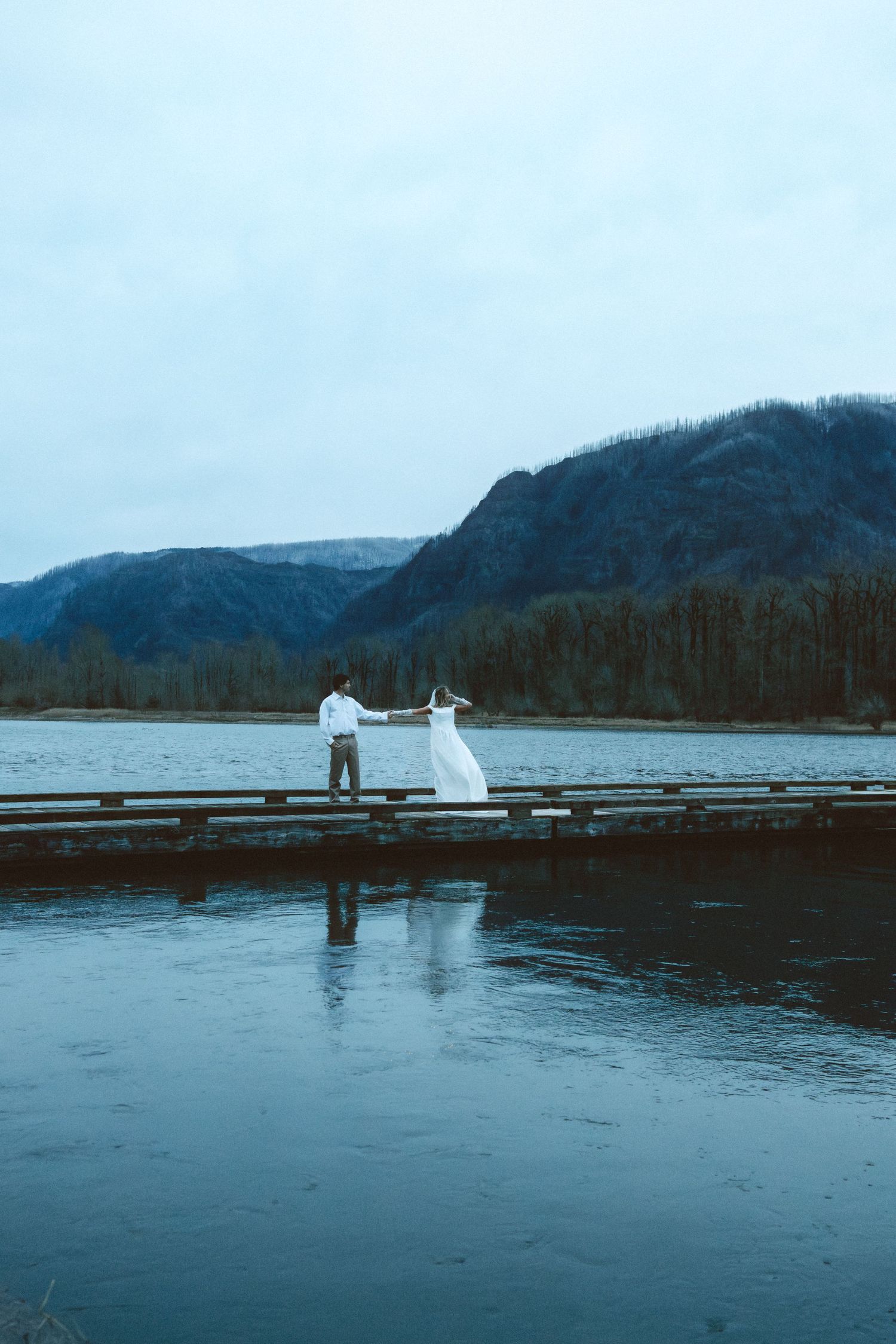 A couple in wedding attire stands on a wooden dock overlooking a serene mountain lake on an overcast day.