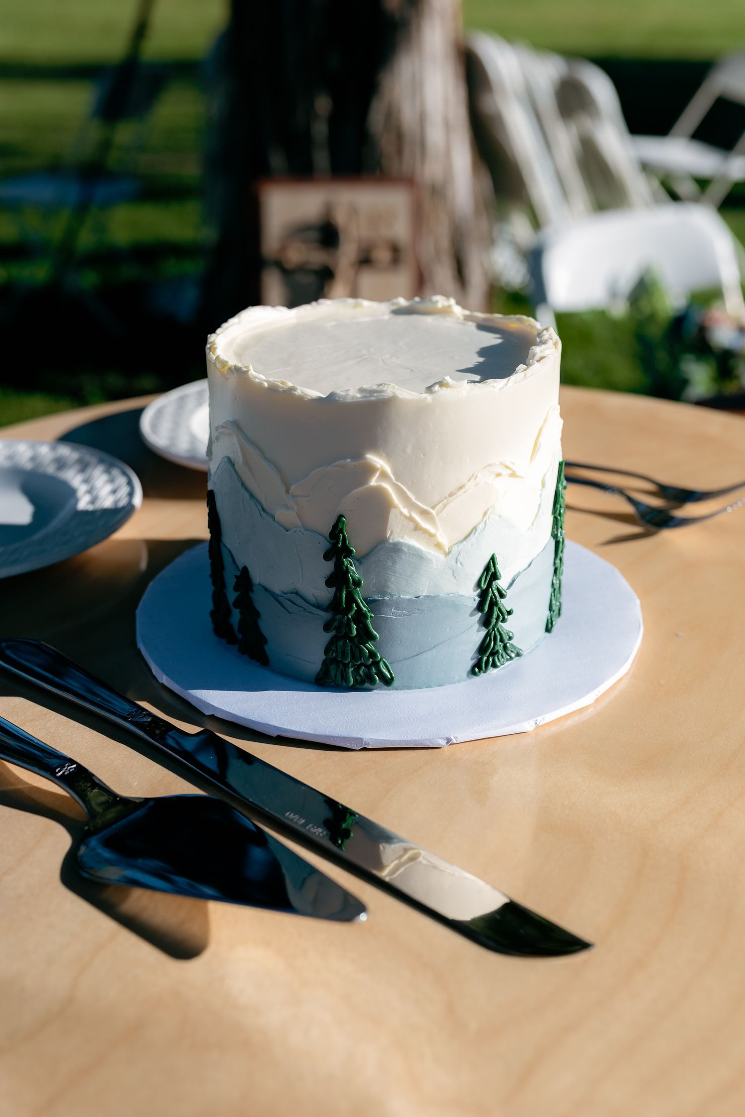 Elegant mountain-themed wedding cake with green pine trees and ombre frosting, displayed on wooden table with serving utensils.