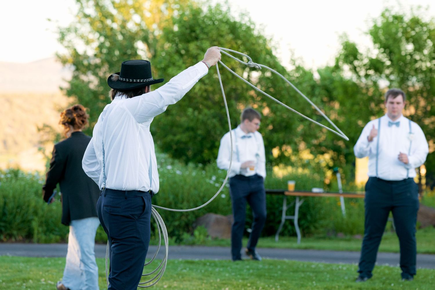 Man in black hat performing lasso rope trick at outdoor wedding reception on lawn.