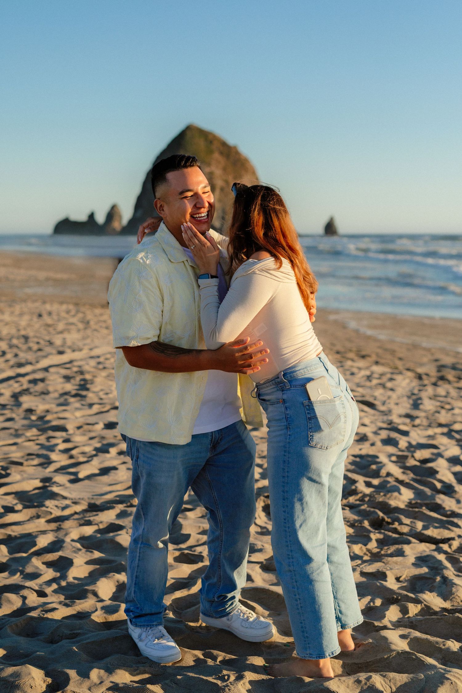 Happy newly engaged couple embracing and laughing on Cannon Beach with Haystack Rock behind them.