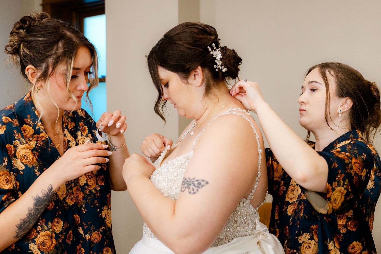 Bridesmaids in floral robes help bride with jewelry during wedding preparations.