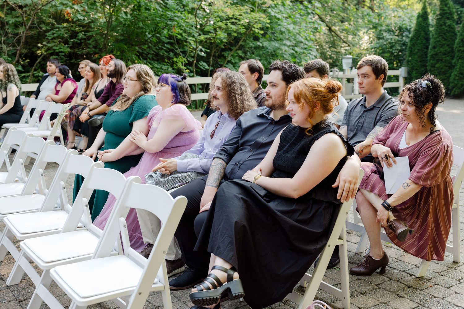 Wedding guests seated in white folding chairs enjoy an outdoor garden ceremony.