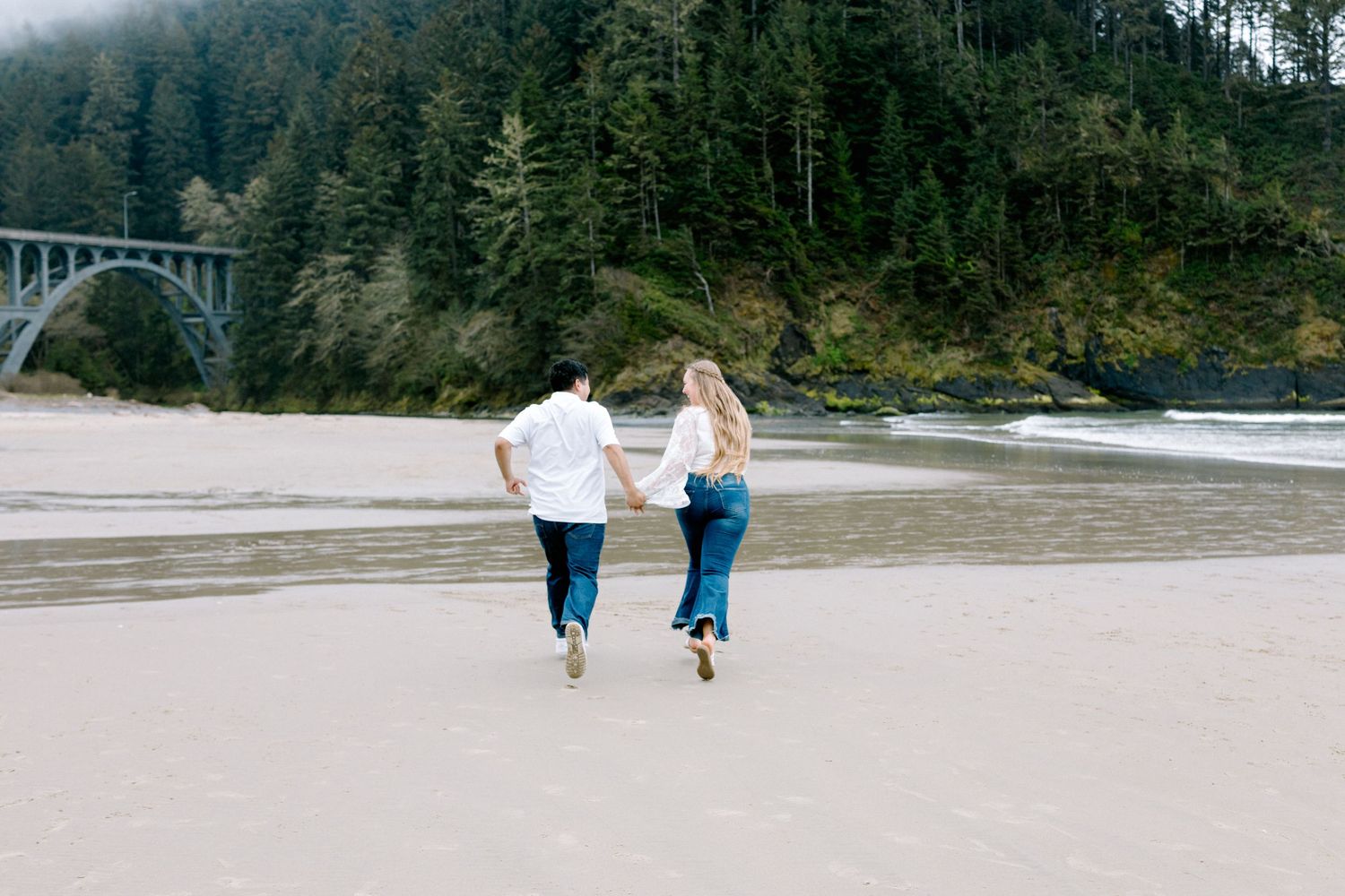Couple running hand in hand on a sandy beach with a bridge and dense forest visible in the background.