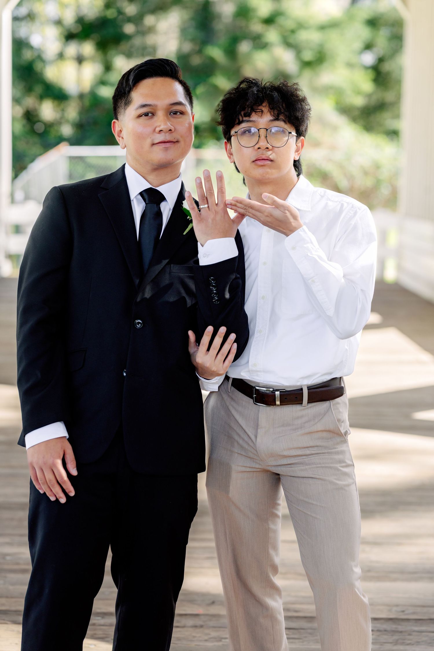 Two men in formal attire display wedding rings, celebrating their marriage outdoors.