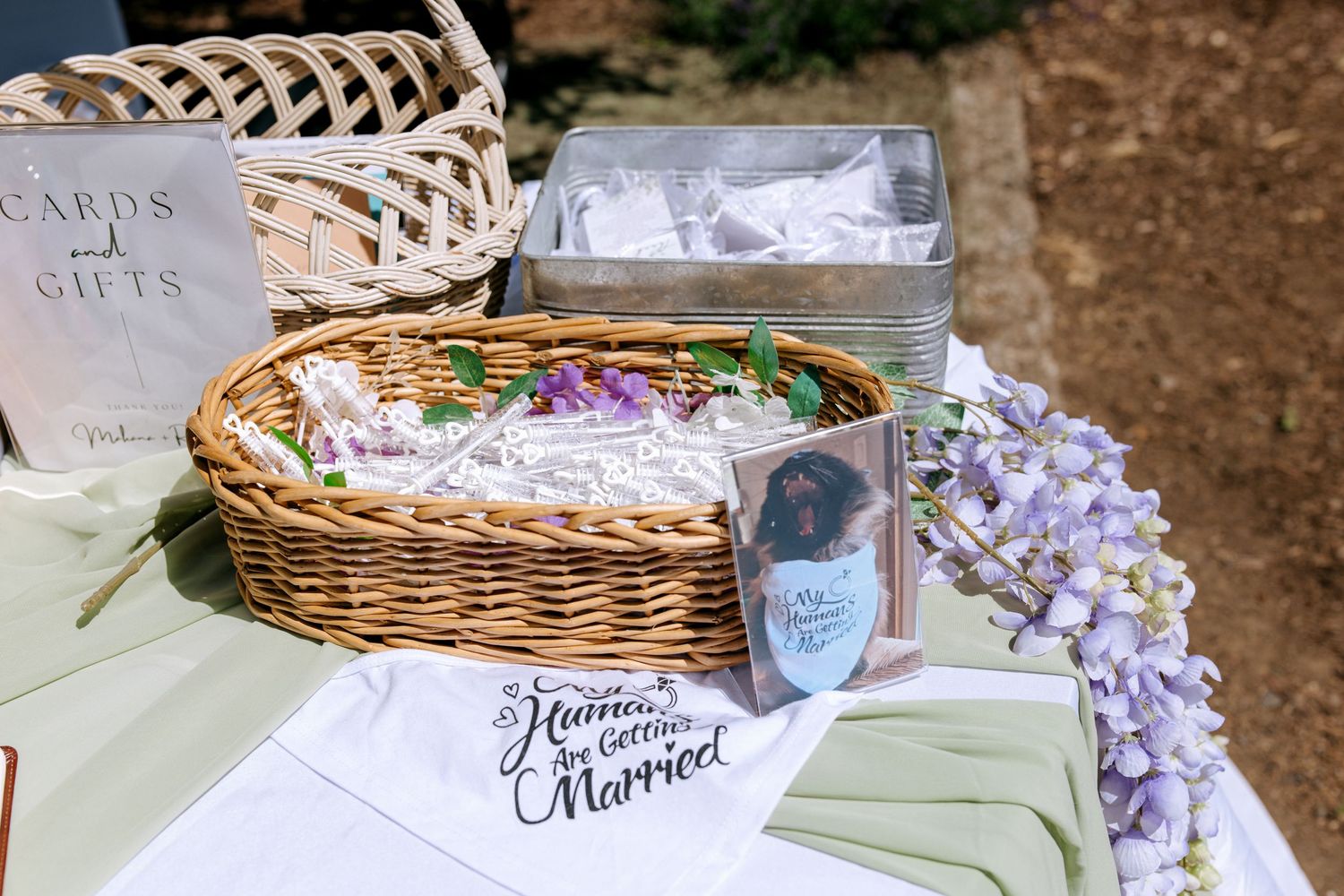 Outdoor wedding gift table with wicker basket, cards, favors, and a decorative “Humans Getting Married” banner.