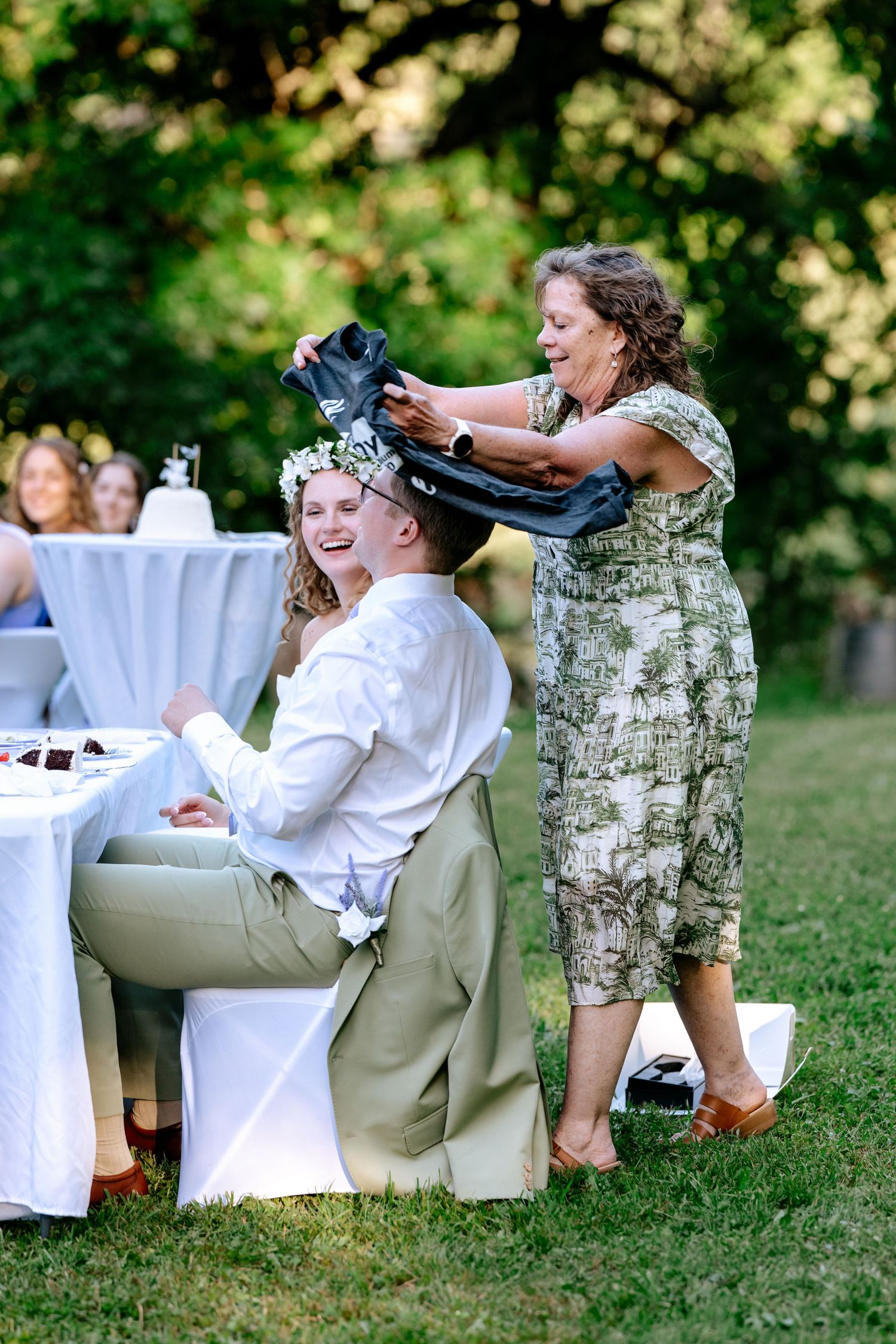 Woman placing jacket over groom's shoulders at outdoor wedding reception on grass.