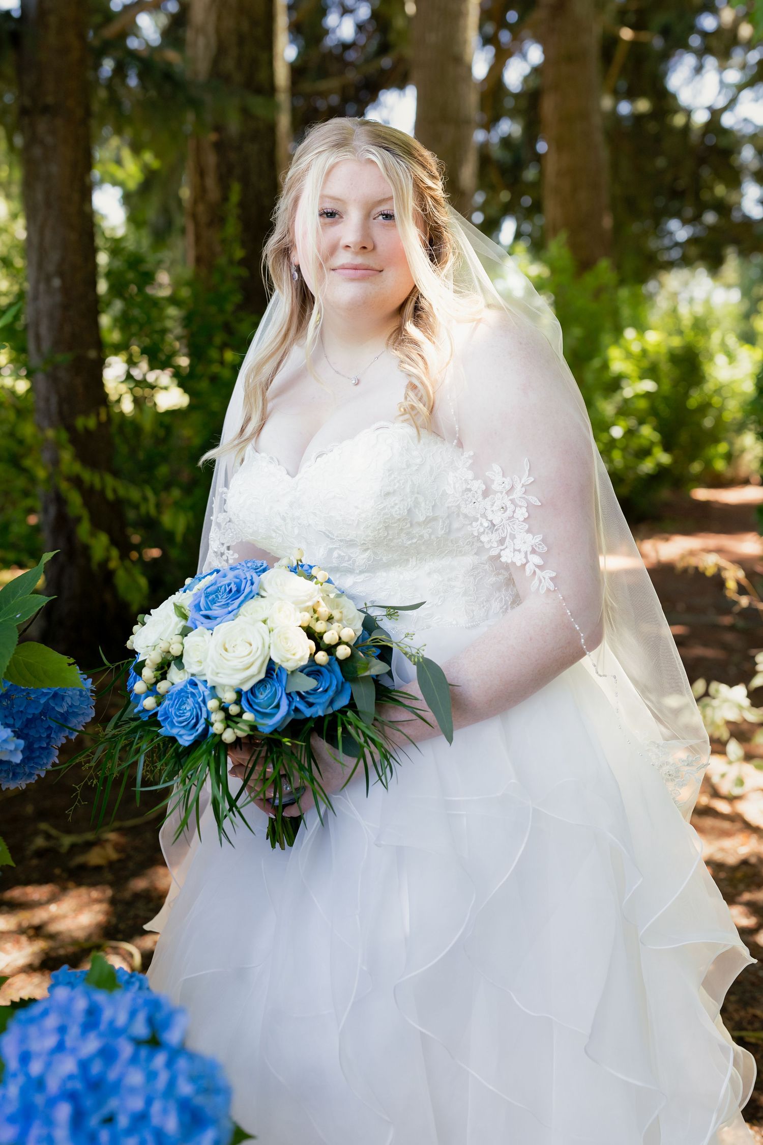 Blonde bride in a white lace gown holds a bouquet of blue and white roses in a garden setting.