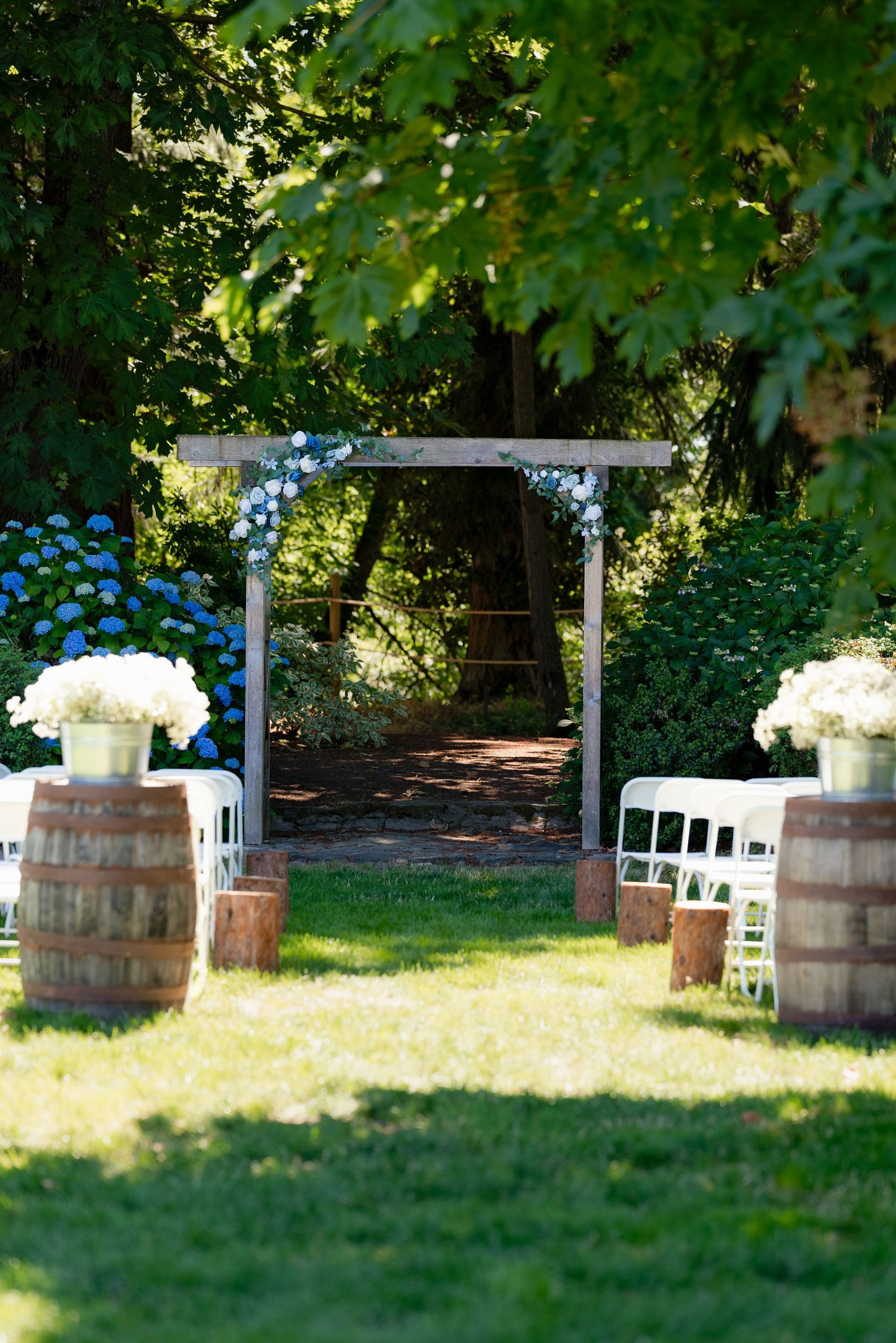 Rustic outdoor wedding ceremony setup with wooden barrel aisle markers, white chairs, and a wooden arch adorned with white flowers.