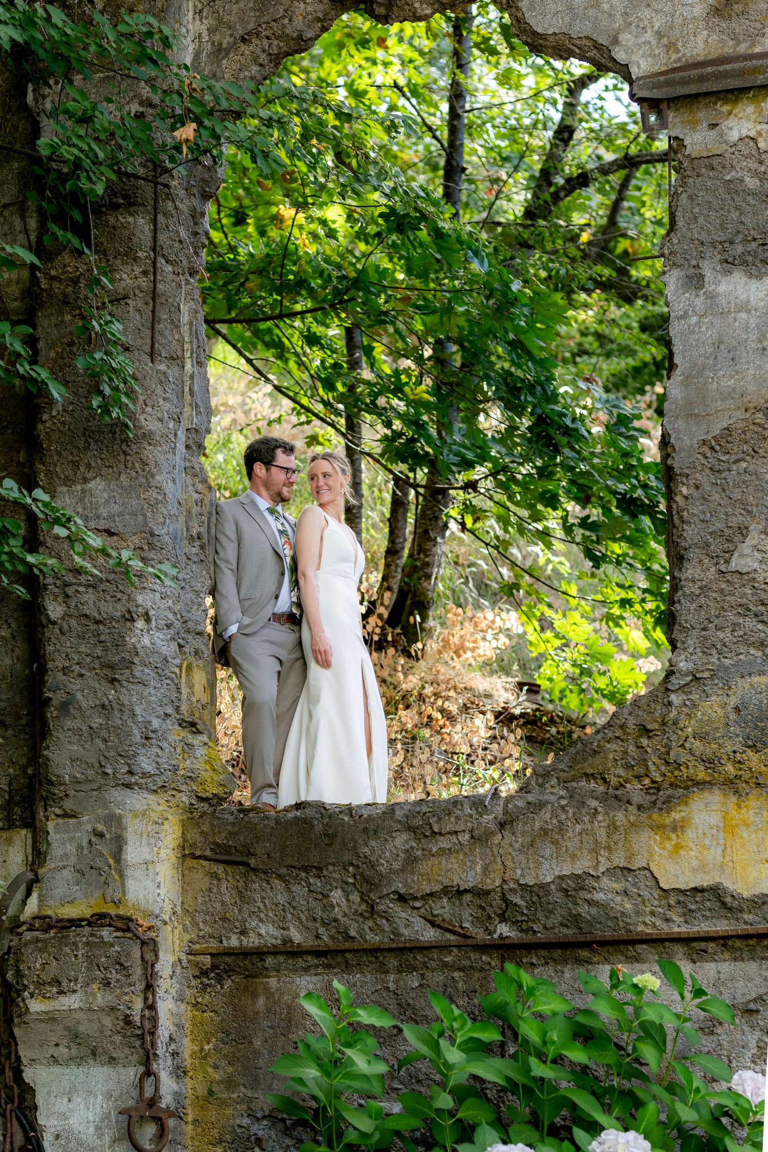 Bride and groom posing in a stone window frame surrounded by lush green trees at an outdoor wedding venue.