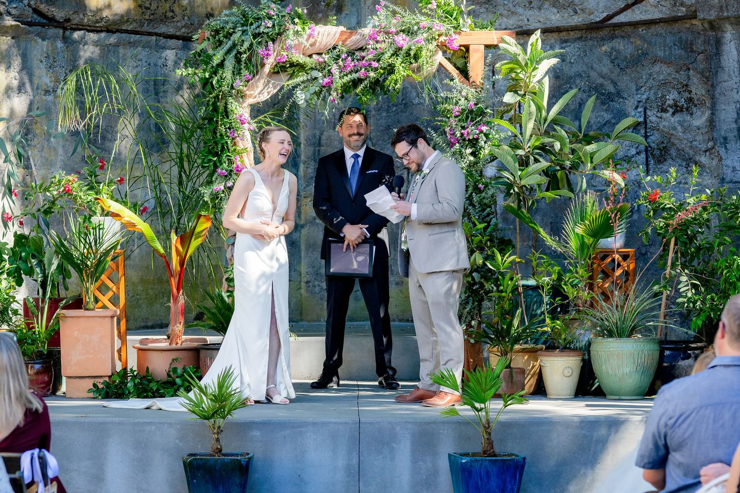 Outdoor wedding ceremony with bride and groom exchanging vows before officiant, surrounded by lush tropical plants and floral arch.