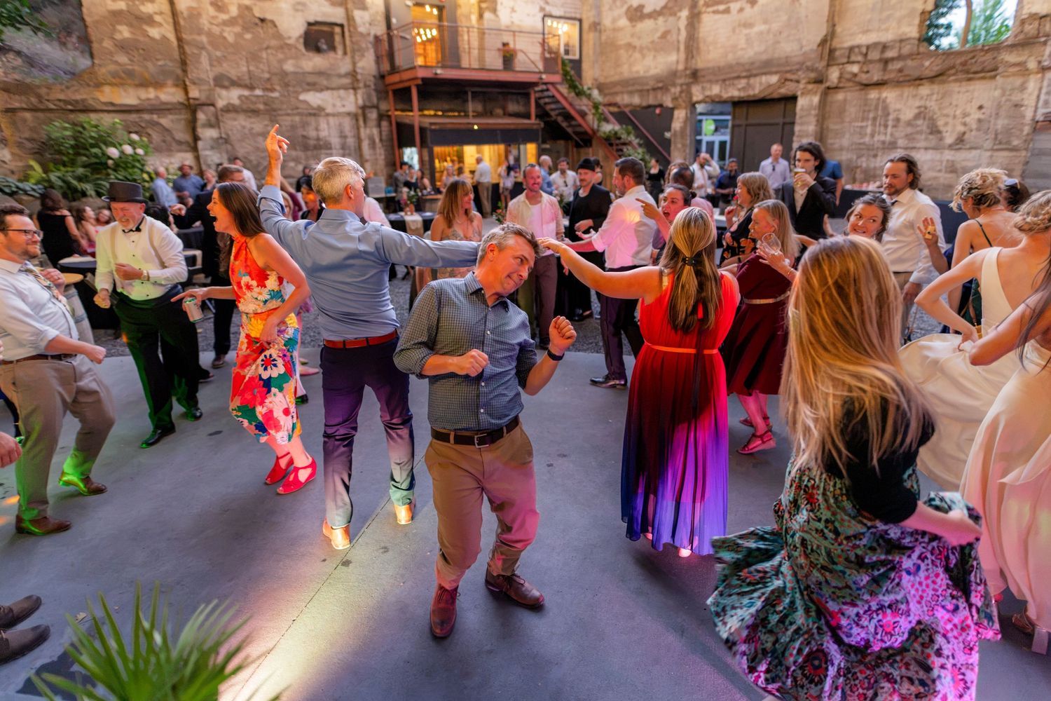 Wedding guests dance energetically together in outdoor courtyard venue illuminated by warm evening lights.