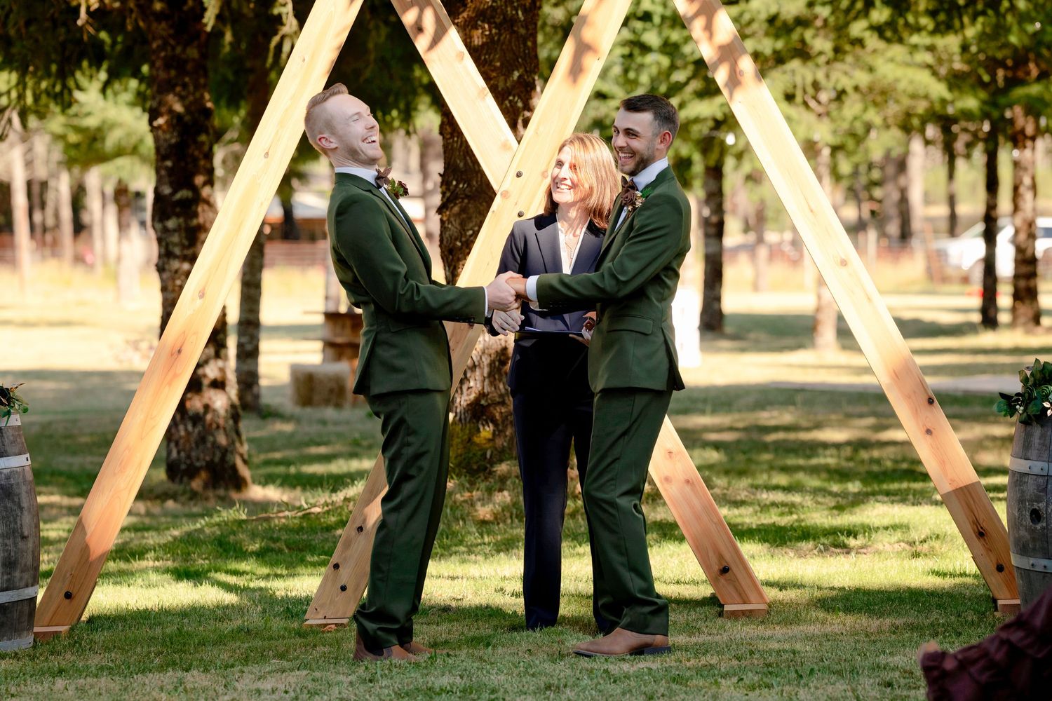 Two grooms in green suits exchange vows under a wooden triangle arch with a female officiant outdoors.