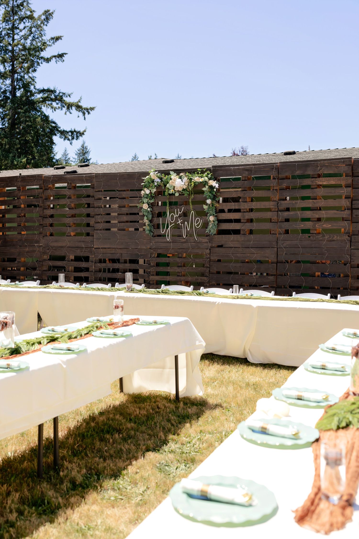 Elegant outdoor wedding reception with long white tables, green place settings, and a wooden pallet backdrop with floral decor.