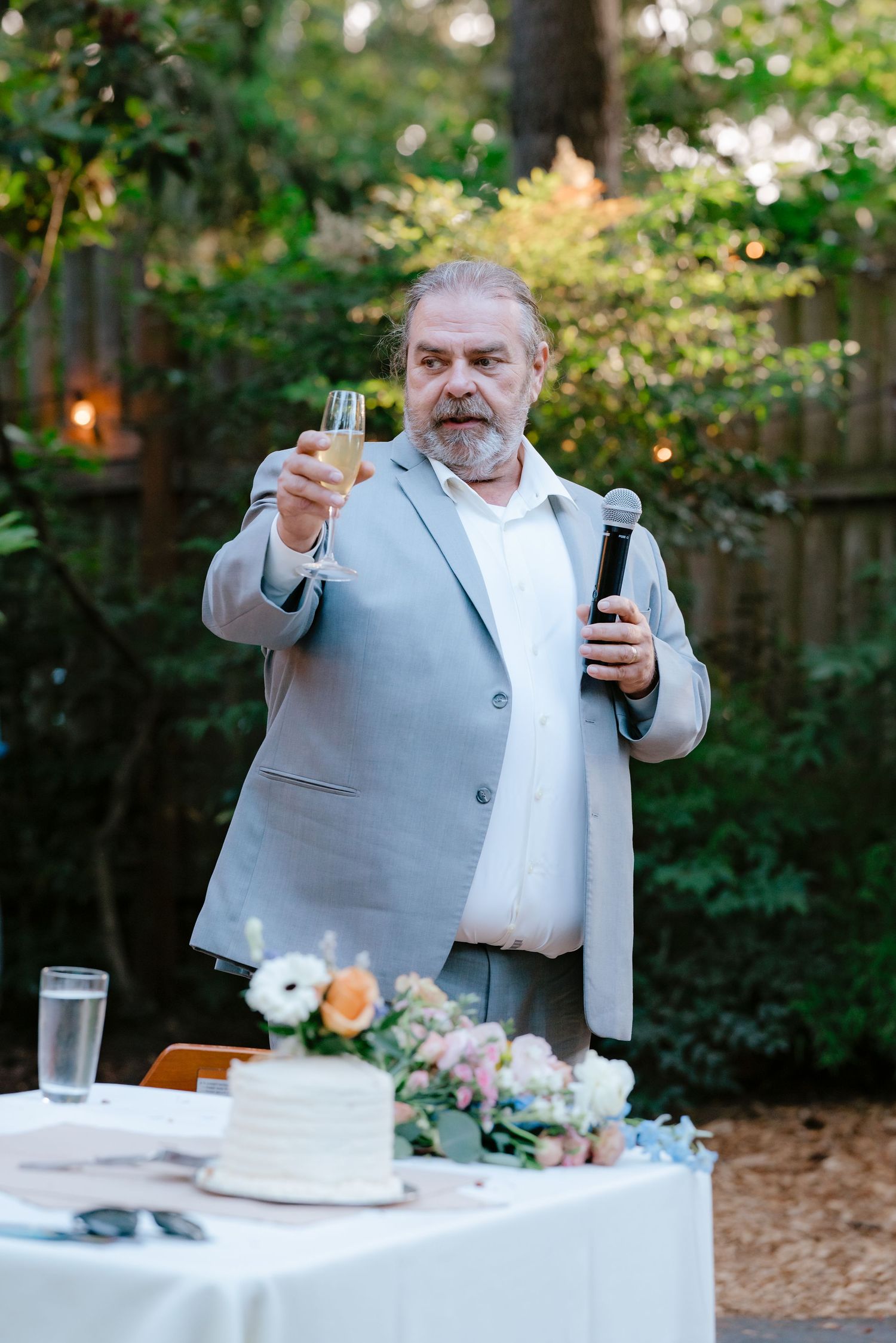 Older man in gray suit raises champagne glass while giving toast at outdoor wedding reception.