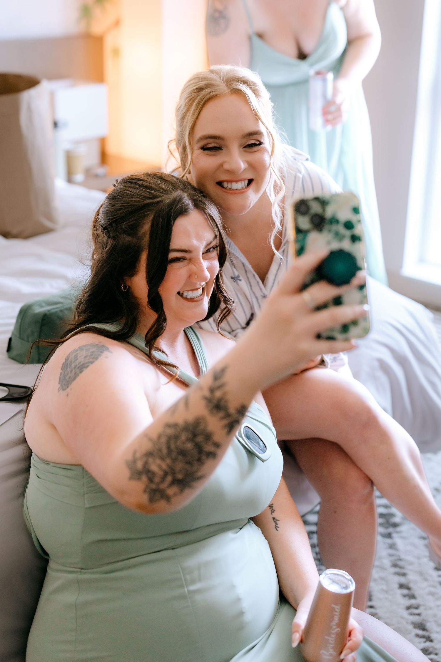 Two bridesmaids in sage green dresses laugh while taking a selfie during wedding preparations.