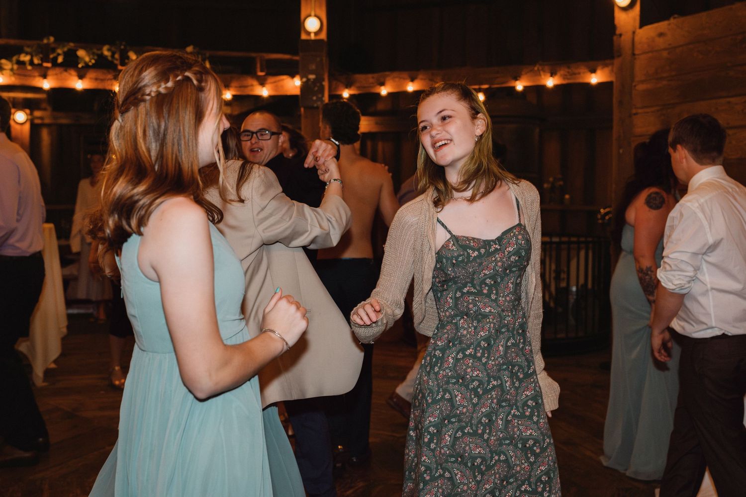 Two young women laughing and dancing together at wedding reception on crowded dance floor.