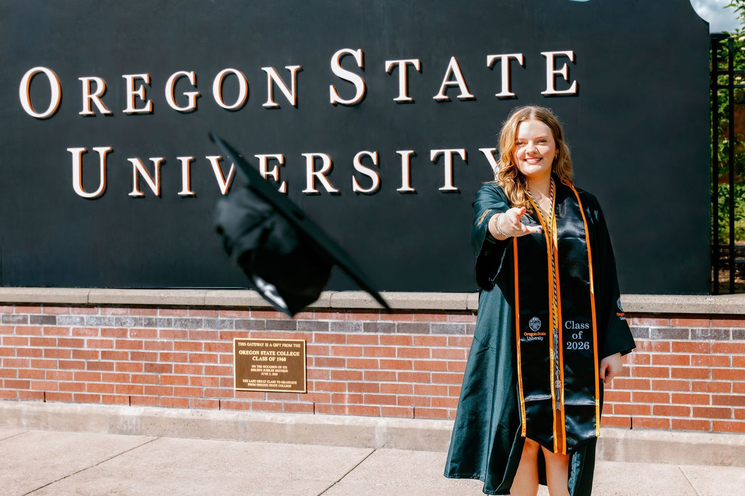 Oregon State University graduate in green cap and gown smiling in front of the OSU brick sign on campus.