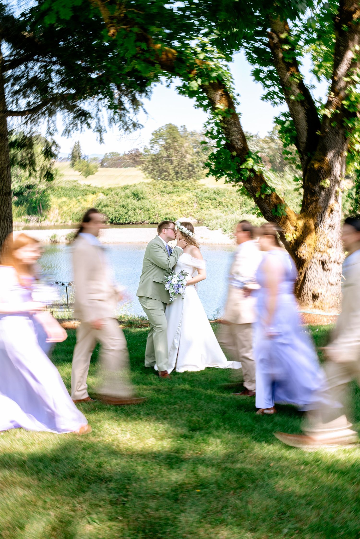 Bride and groom share a kiss outdoors near a lake, surrounded by blurred wedding party in lavender and tan attire.