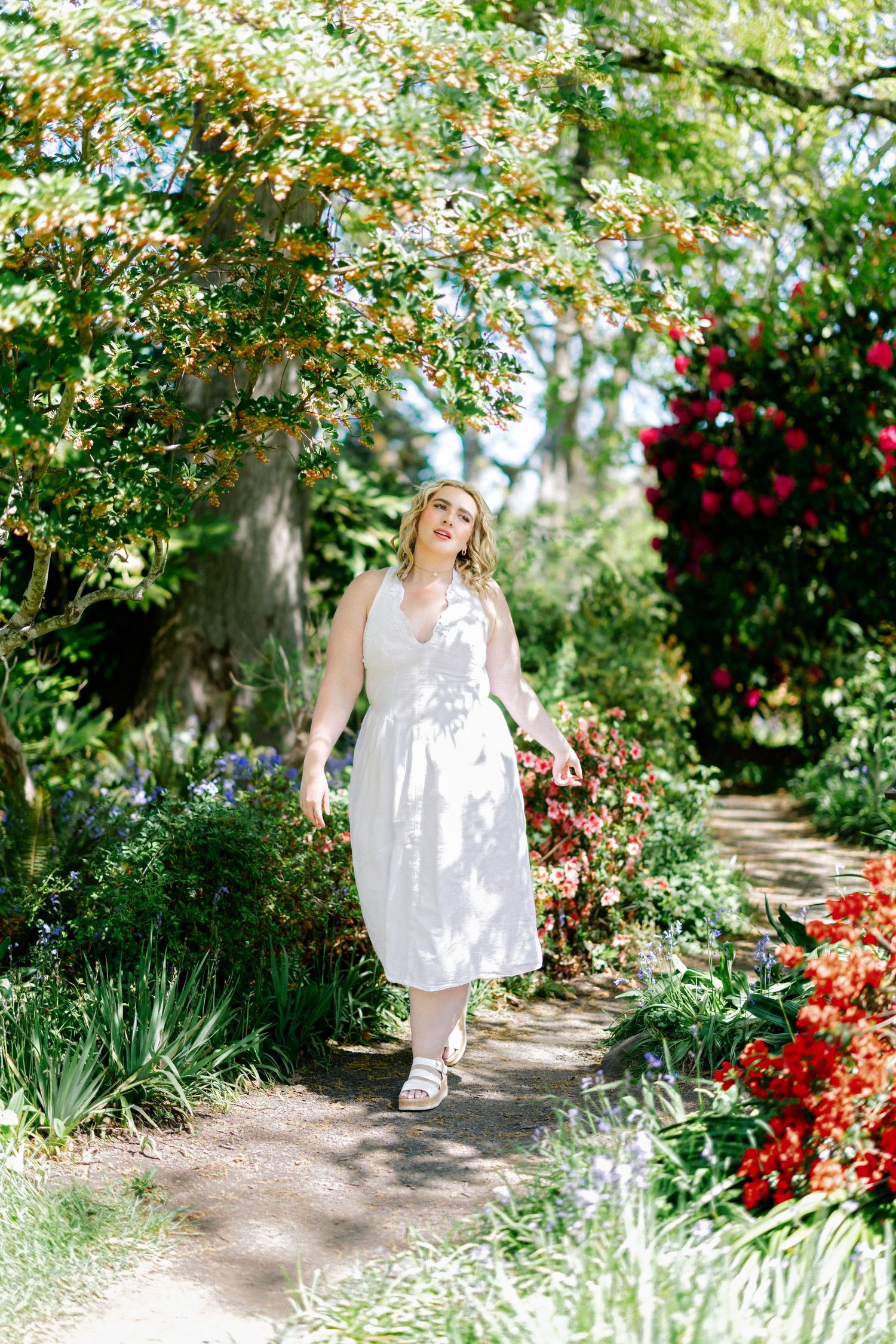 Woman in white dress walking along a garden path surrounded by colorful flowering shrubs.