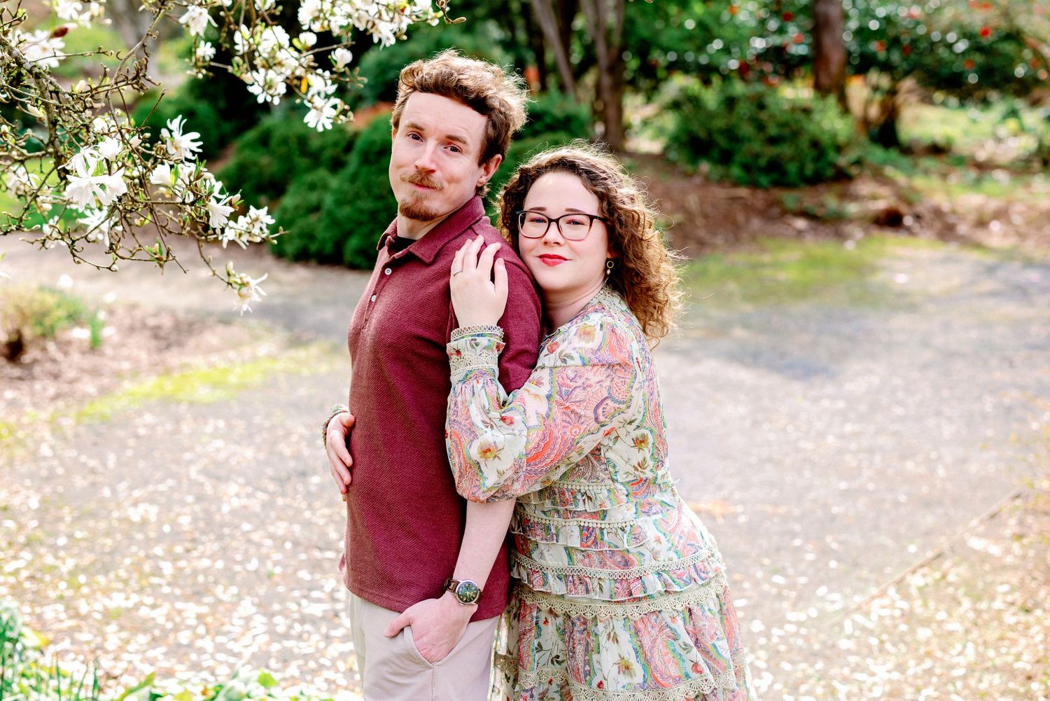 A couple embraces lovingly outdoors surrounded by blooming spring trees and scattered flower petals on the ground.