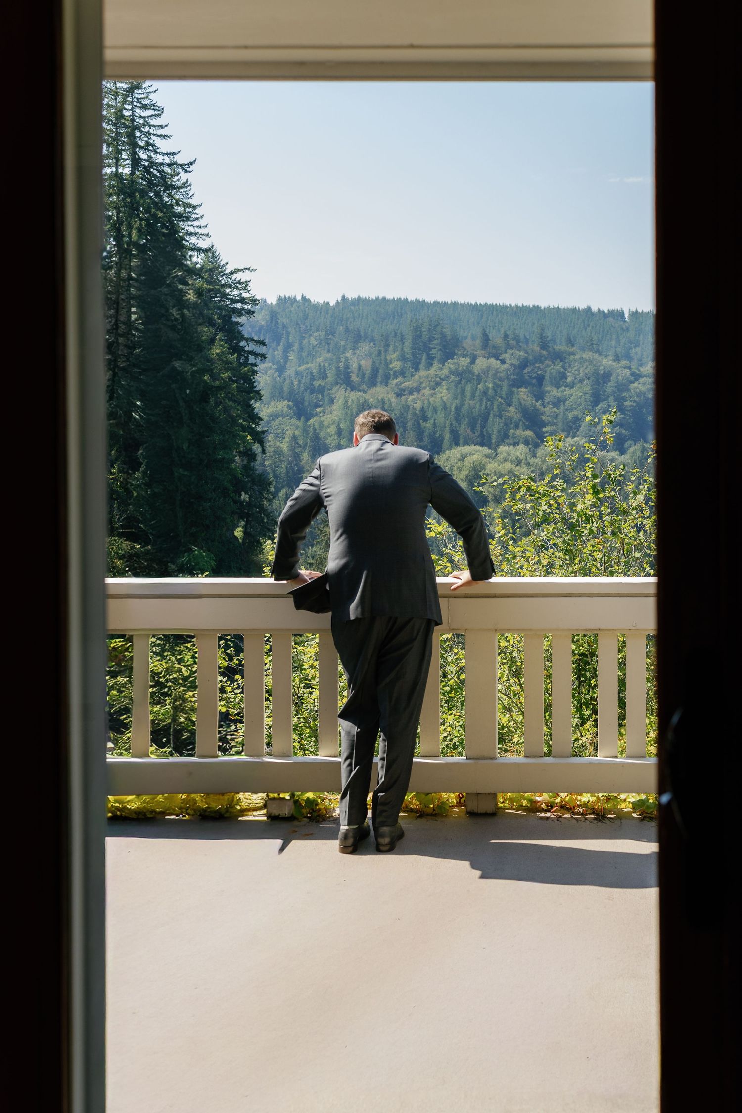 A person in a suit stands at a balcony railing overlooking a forested mountain landscape on a sunny day.