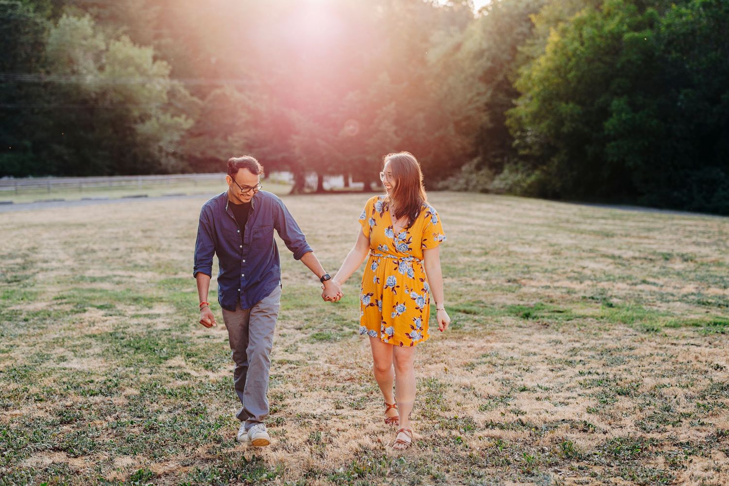 A couple holds hands while walking through a sun-drenched field at golden hour in the countryside.