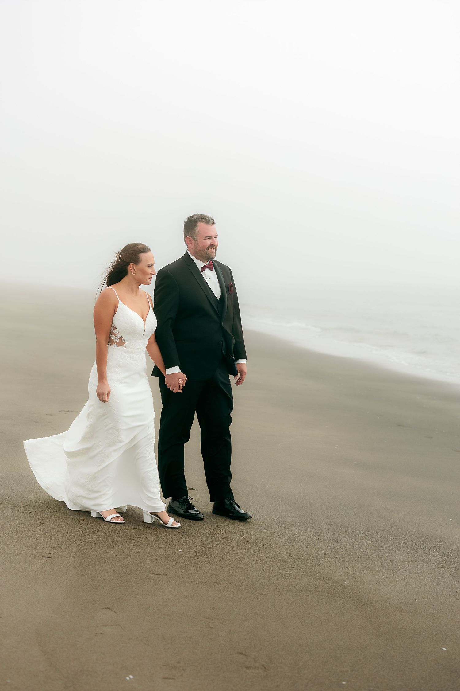 Wedding couple walking along a misty beach shore in formal attire.