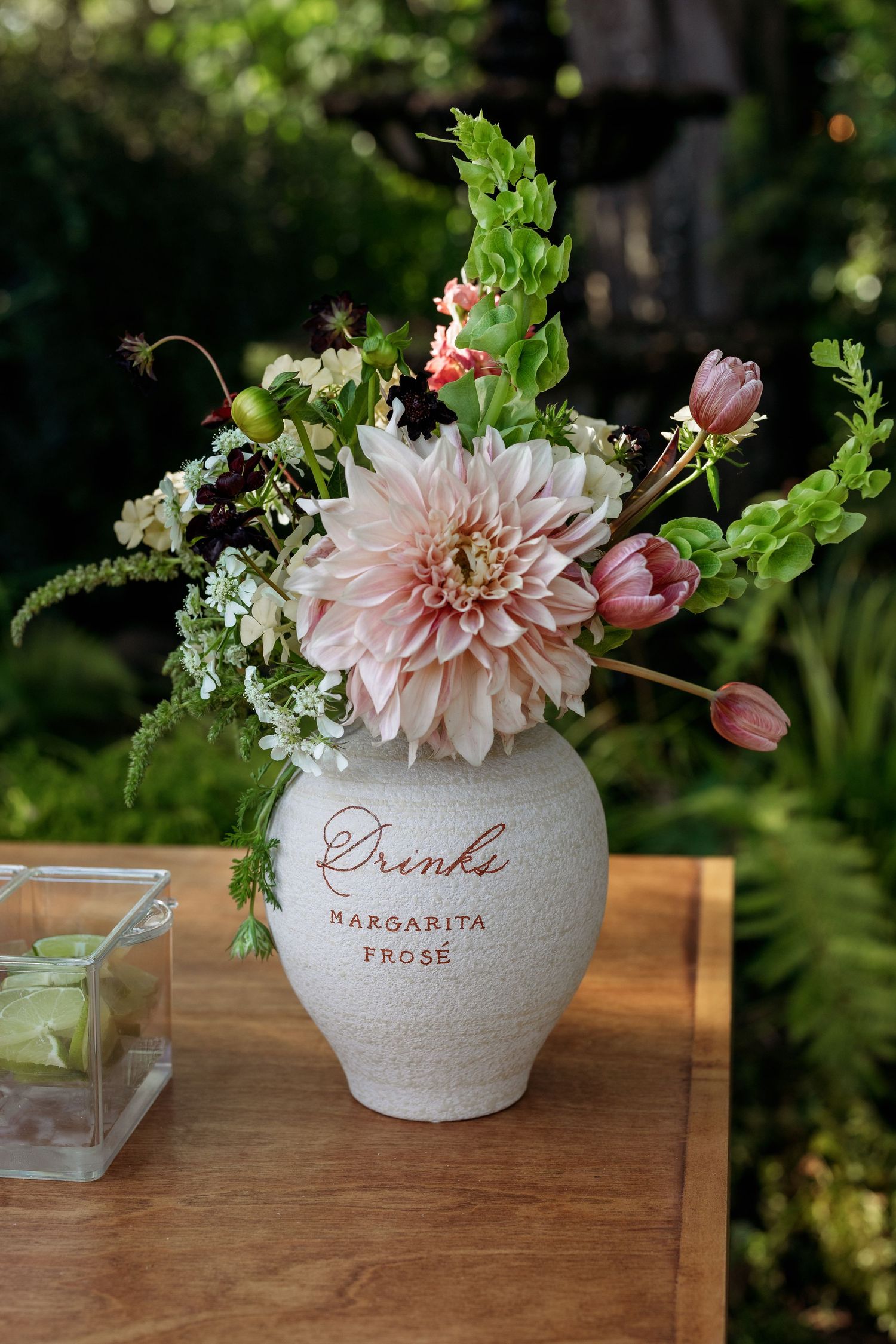 Pink dahlia flowers arranged in white ceramic vases with text on wooden table in garden setting.