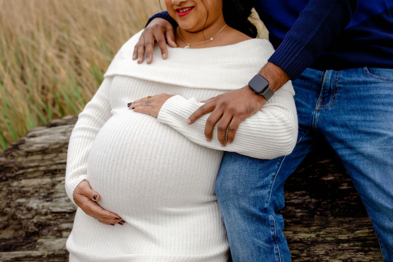 A person in a white sweater cradles their pregnant belly while standing in a grassy outdoor setting.