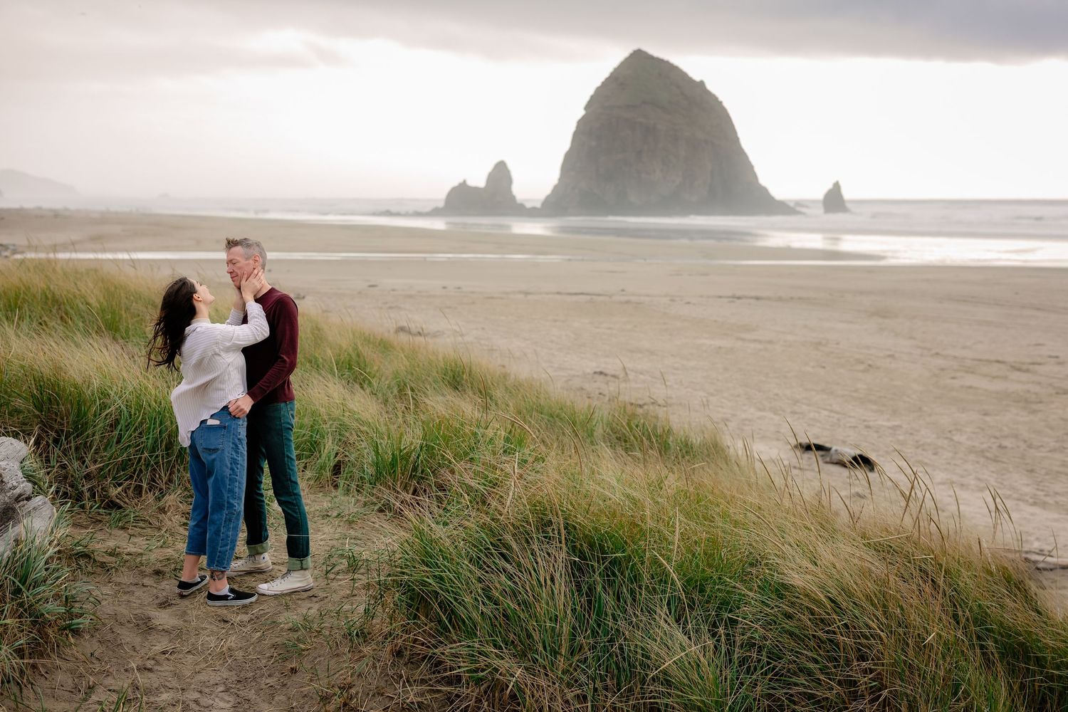 A couple stands on grassy dunes overlooking Cannon Beach with iconic Haystack Rock visible in the misty distance.