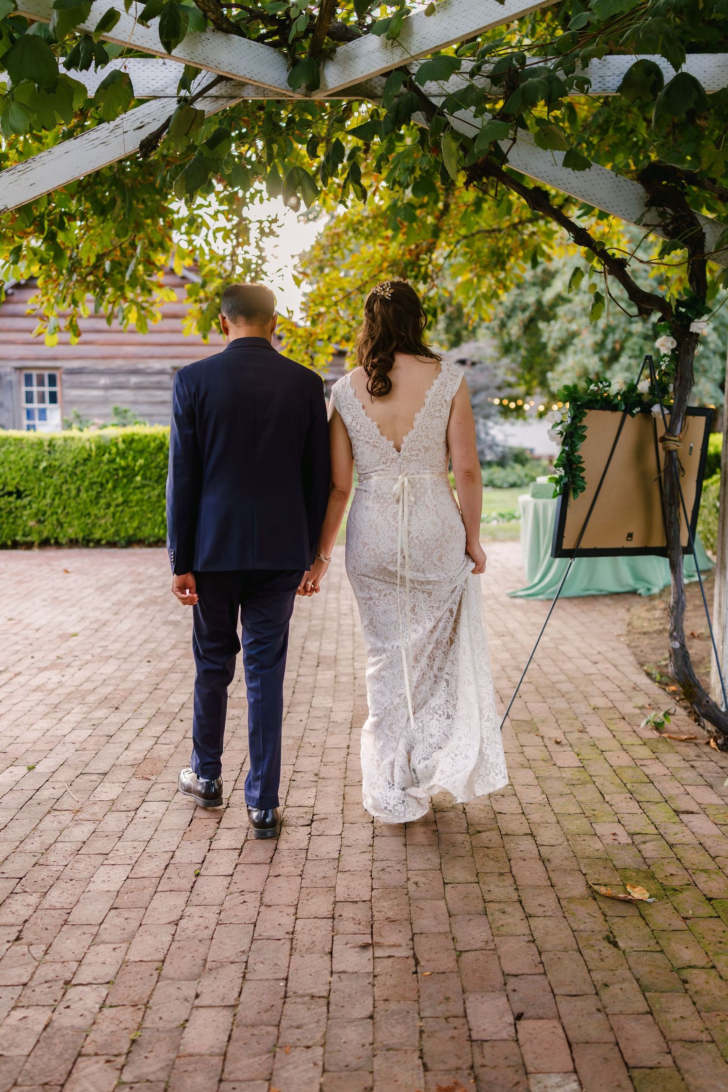 A couple walks along a brick pathway under green leafy vines on their wedding day.