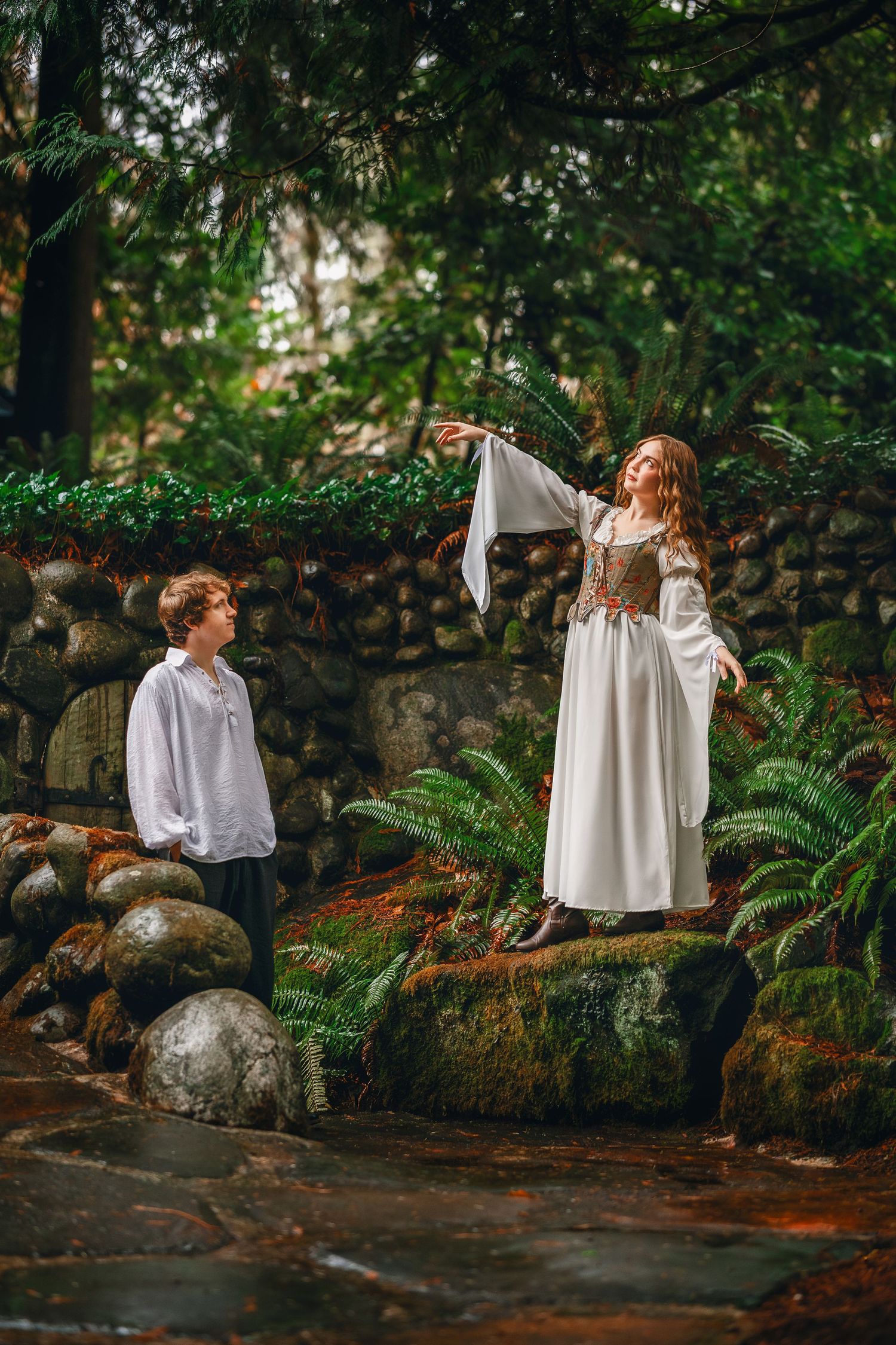 Two figures in white flowing garments dance among mossy rocks in an enchanted forest setting.