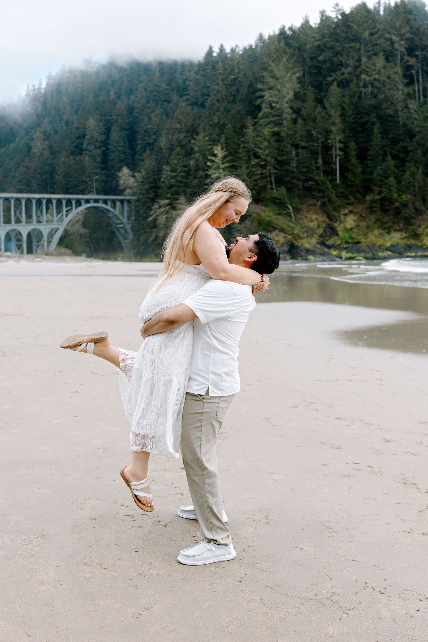 Man lifting woman in white dress on Oregon coast beach with bridge and forest backdrop.