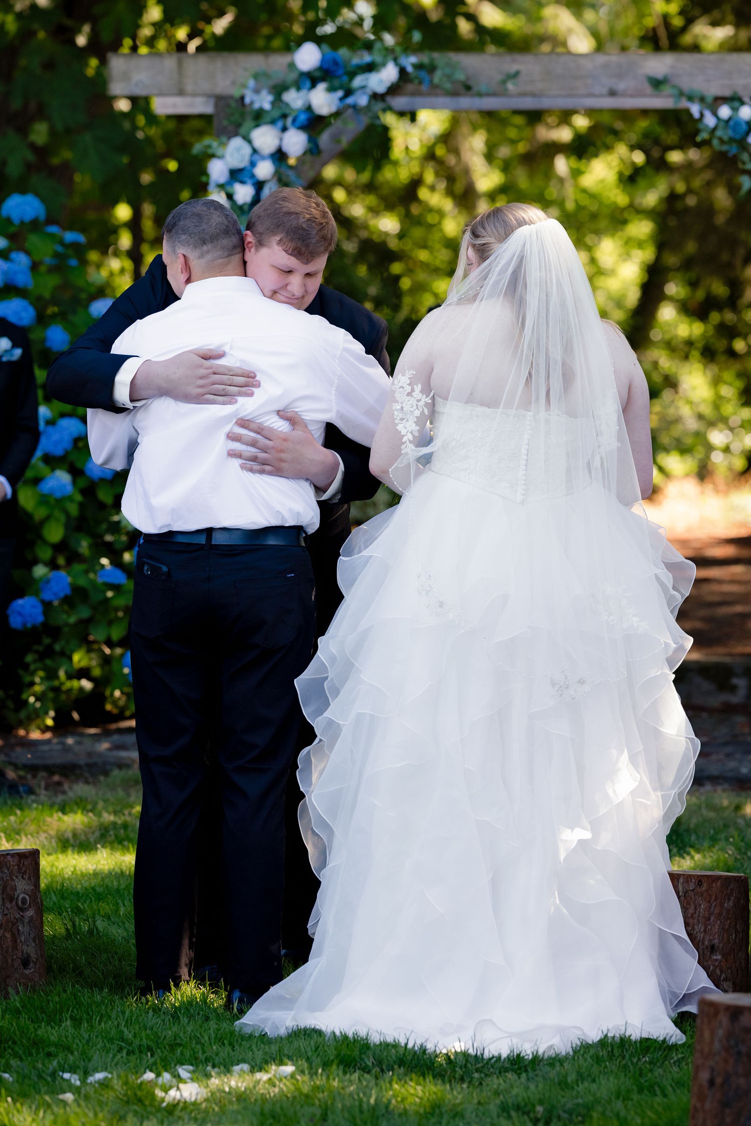 Emotional wedding ceremony moment as groom hugs family member at outdoor altar with blue hydrangeas.