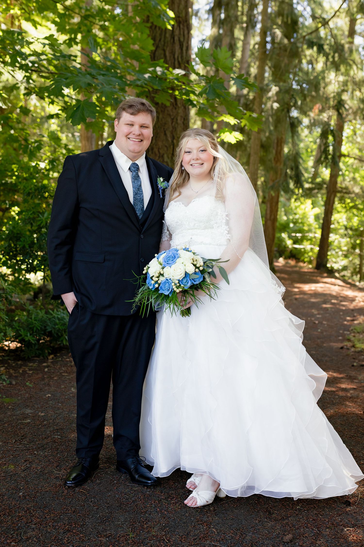 Smiling bride and groom pose together outdoors holding blue and white wedding bouquet.