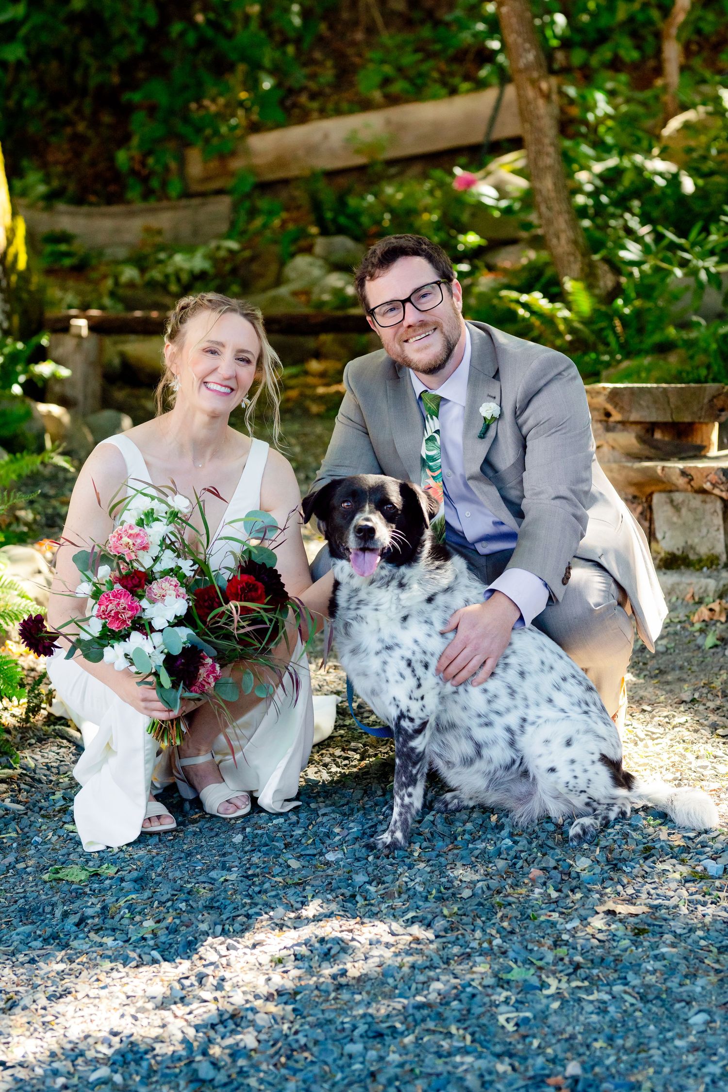 Smiling bride and groom pose with their spotted dog on gravel path surrounded by lush greenery on wedding day.