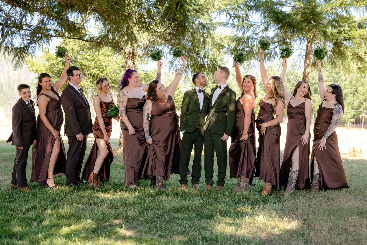 Wedding party in brown attire celebrates joyfully with raised hands under pine trees on a sunny day.