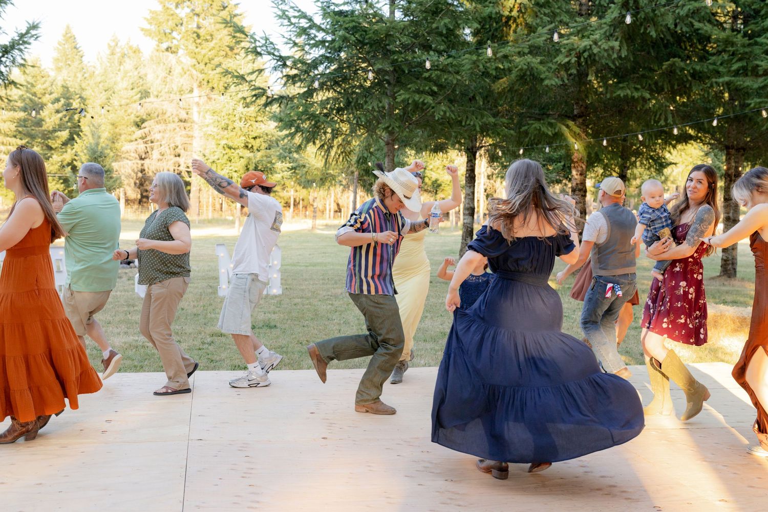 Guests dancing joyfully outdoors at a wedding reception surrounded by tall pine trees in golden evening light.