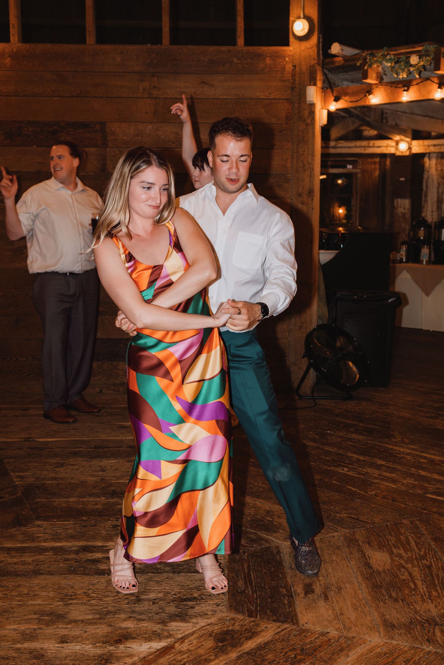 Couple dancing energetically on wooden dance floor at wedding reception, woman in colorful multicolor dress.