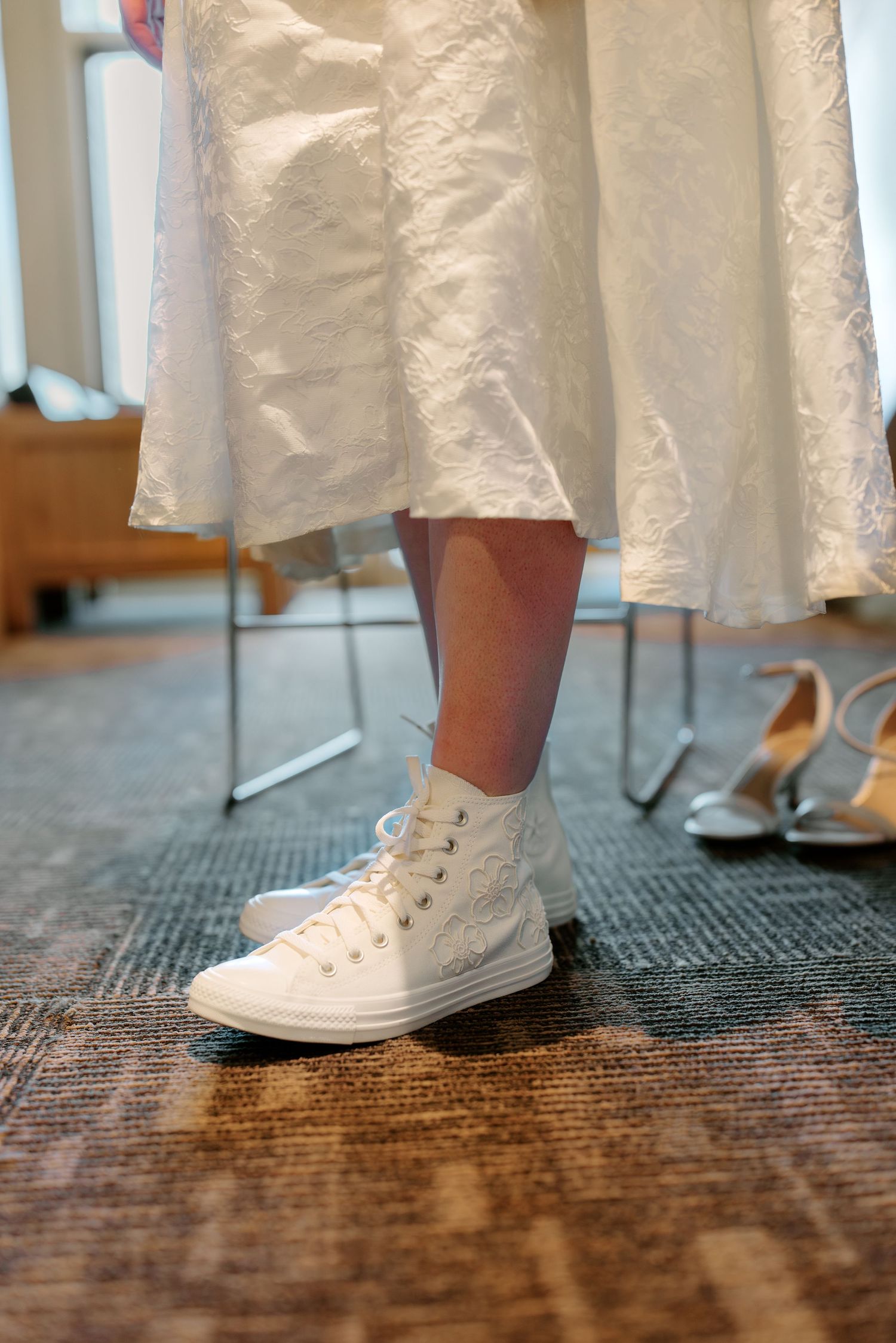 Bride wearing white Converse high-top sneakers under lace wedding dress at wedding venue.
