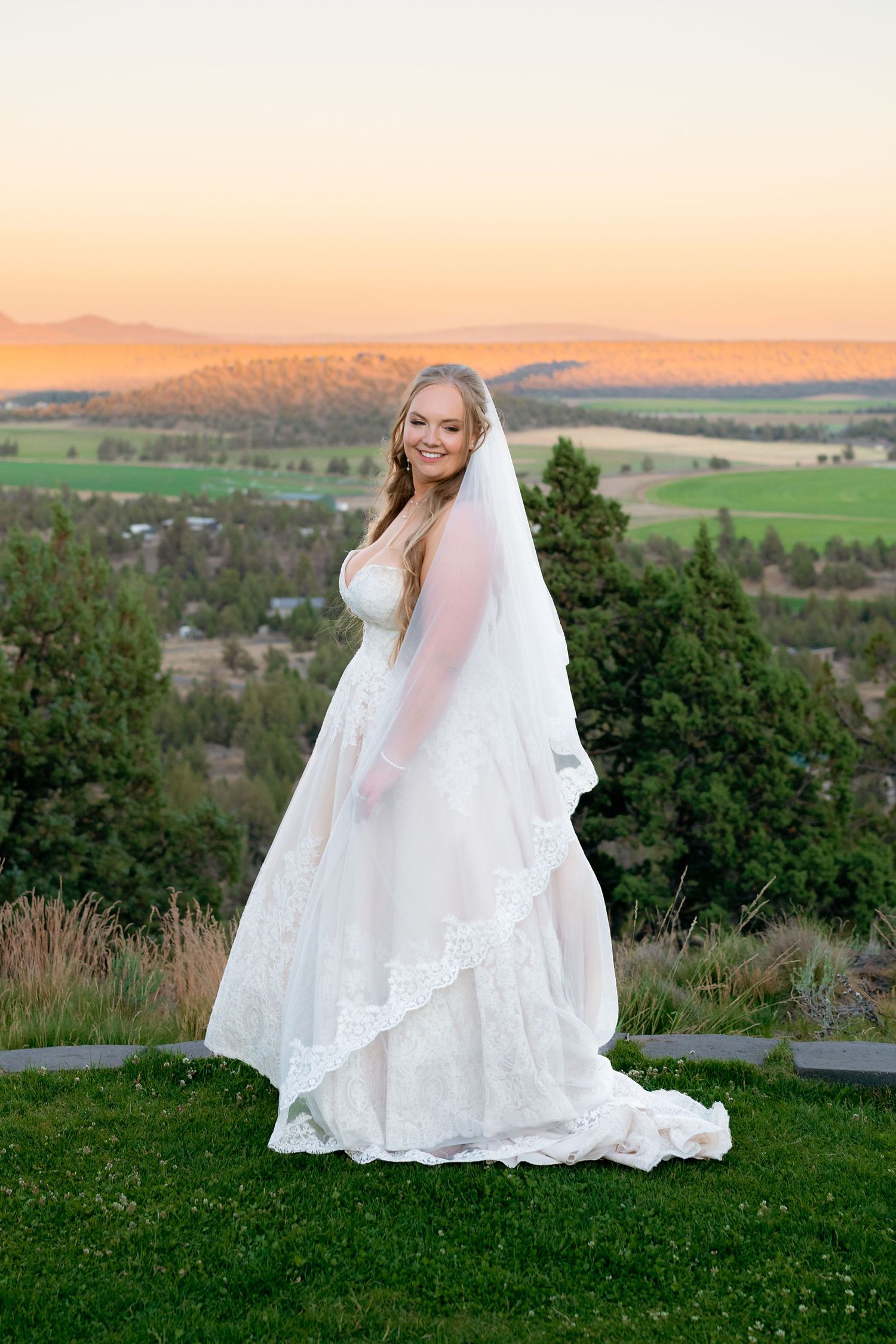 Smiling bride in lace wedding gown and veil poses at sunset with mountain views.
