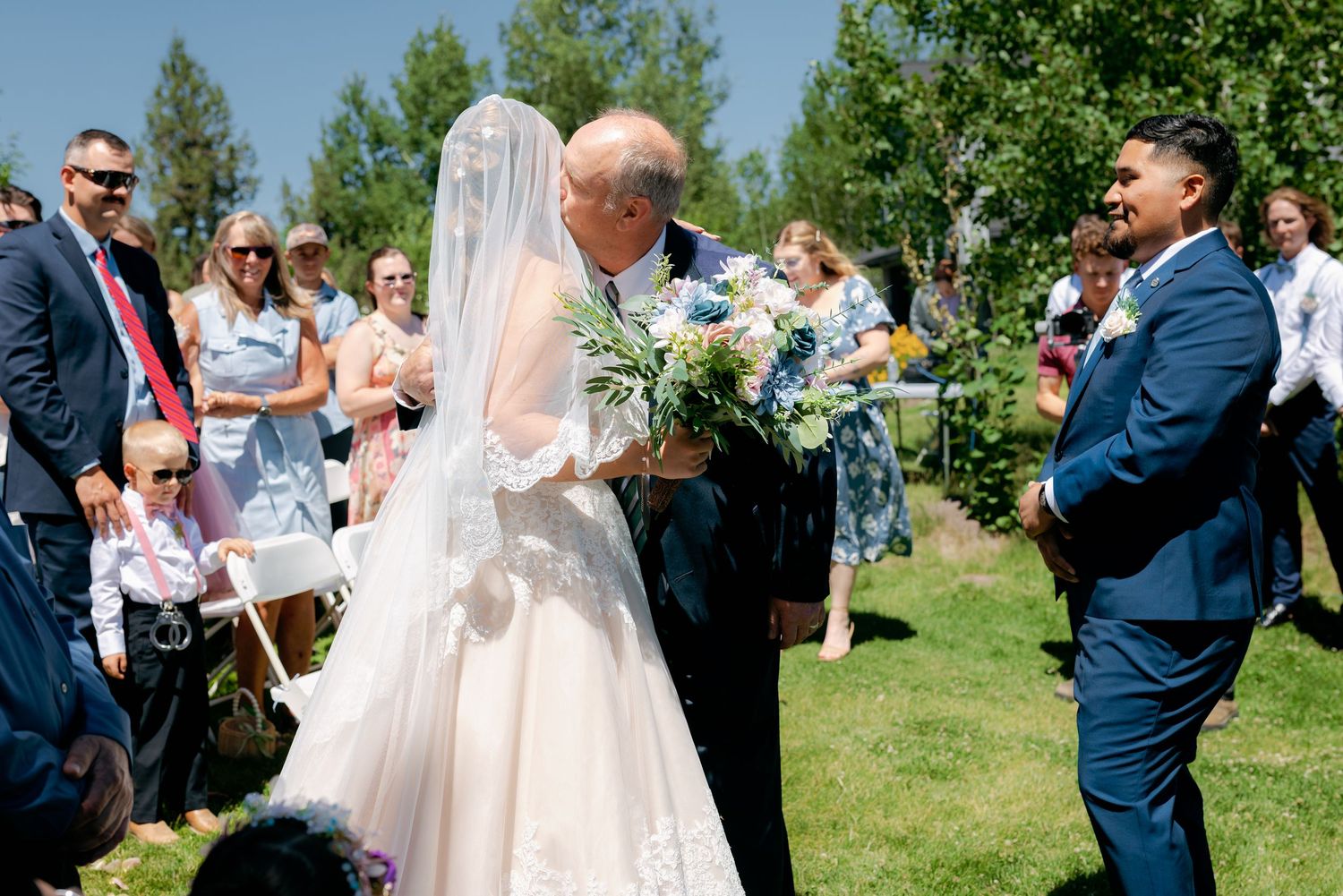 Father of the bride walks daughter down the aisle at sunny outdoor wedding ceremony while groom watches.