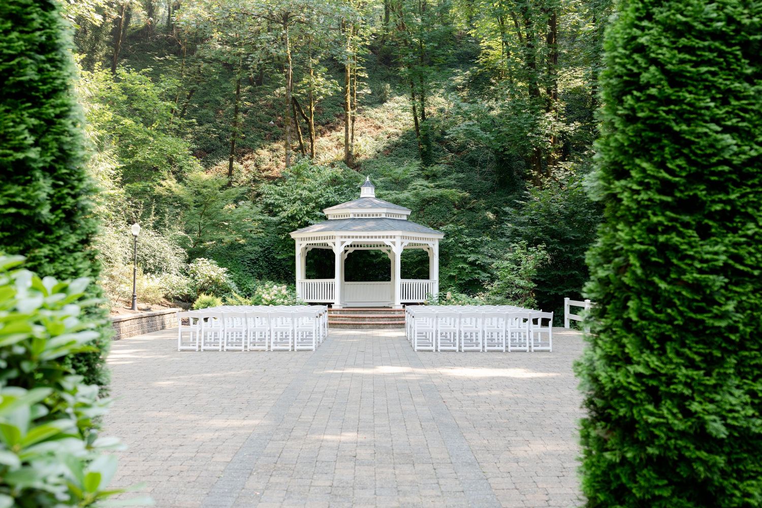 Elegant outdoor wedding ceremony setup with white chairs and a gazebo surrounded by lush green trees.